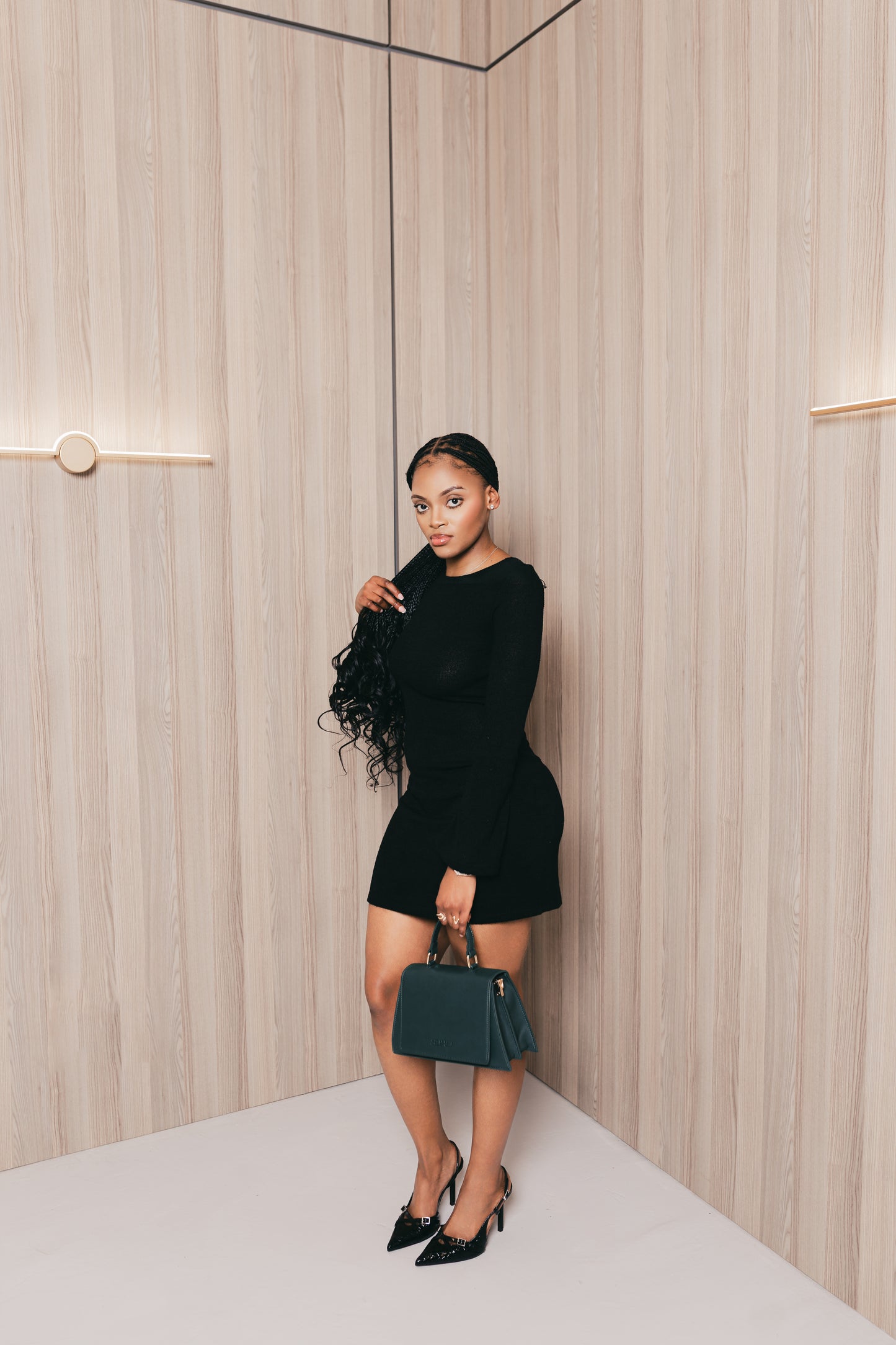 Woman in a black outfit holding a green suhu handbag against a neutral wall.
