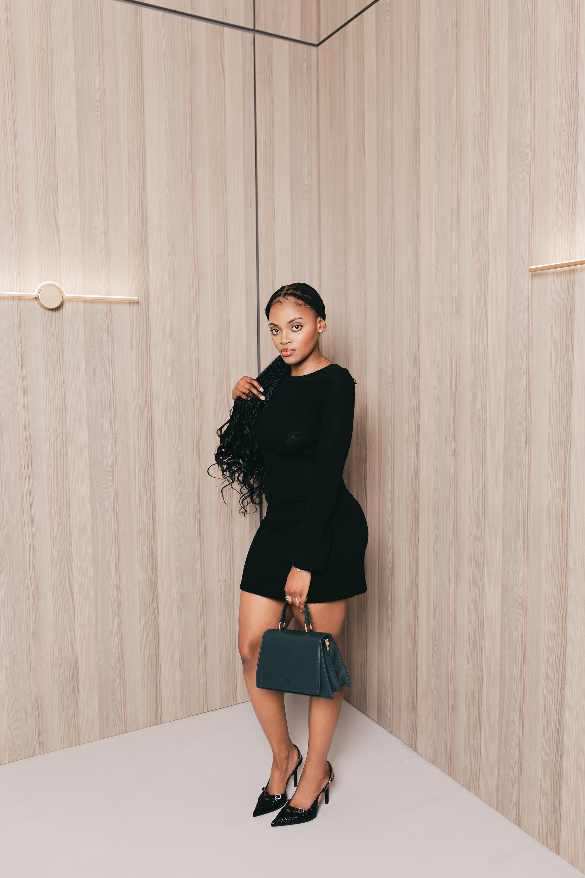 Woman in a black outfit holding a green suhu handbag against a neutral wall.