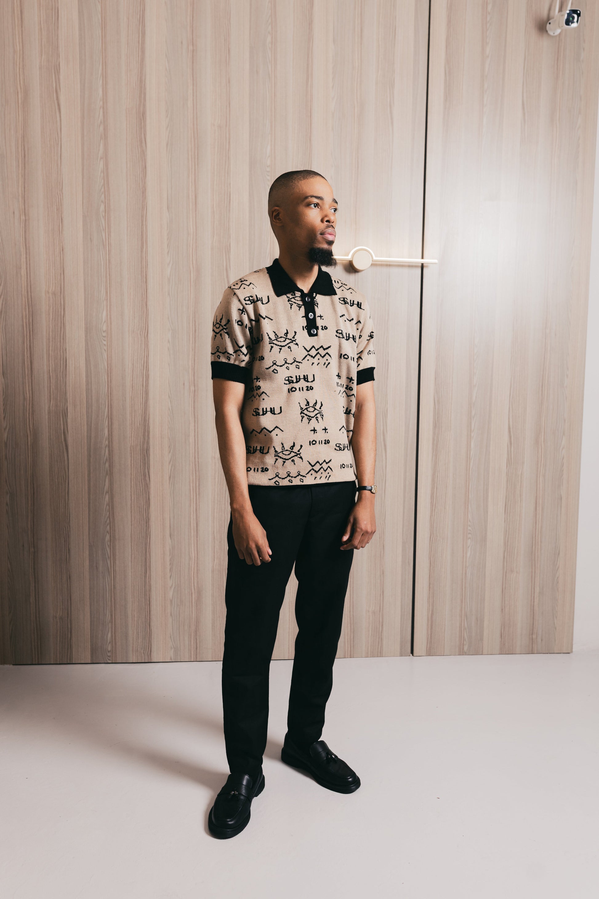 Model wearing a patterned SUHU Golf shirt and black pants standing against a wooden wall.