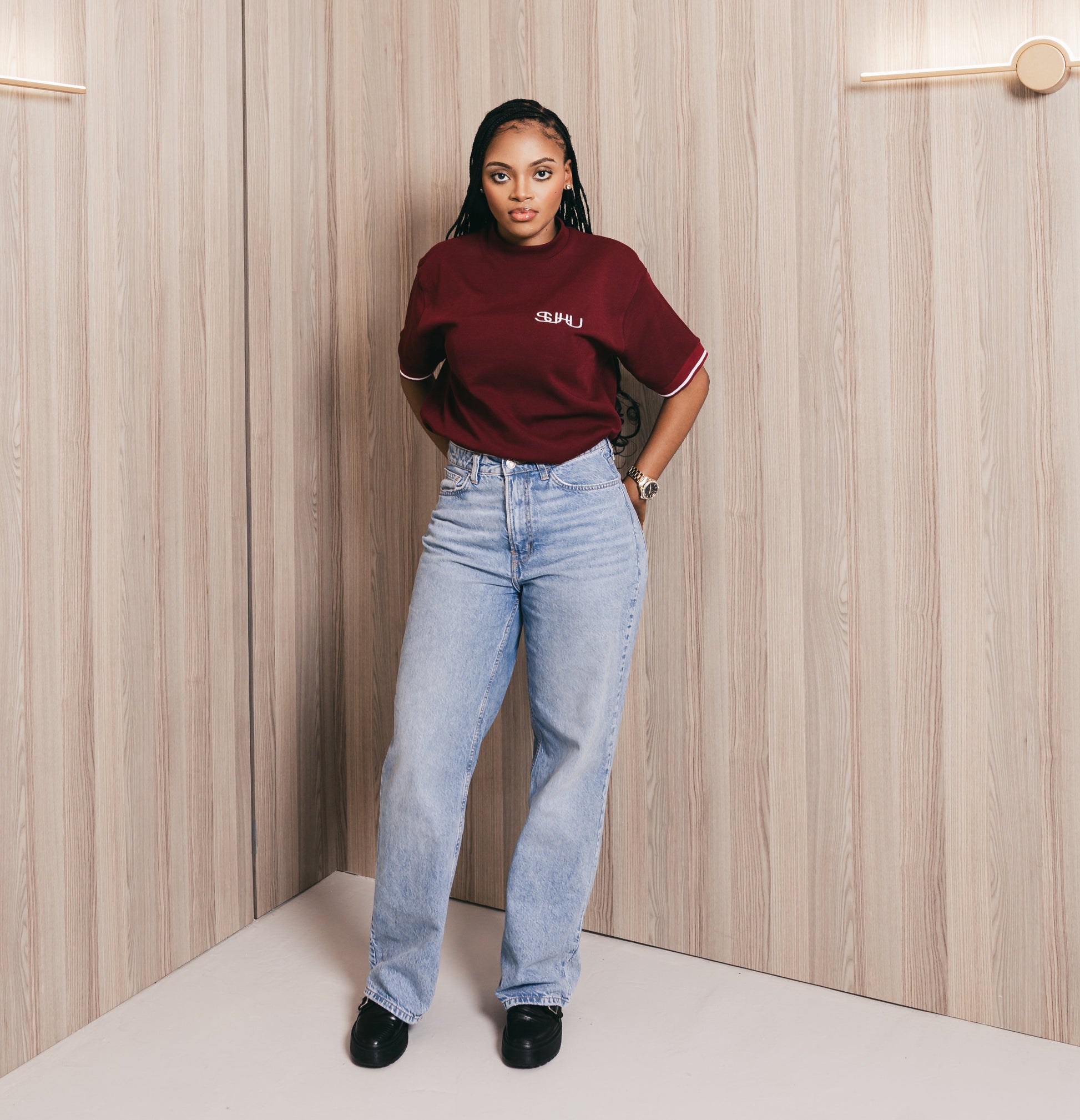 Person wearing a burgundy top and light blue jeans standing against a wooden paneled wall.