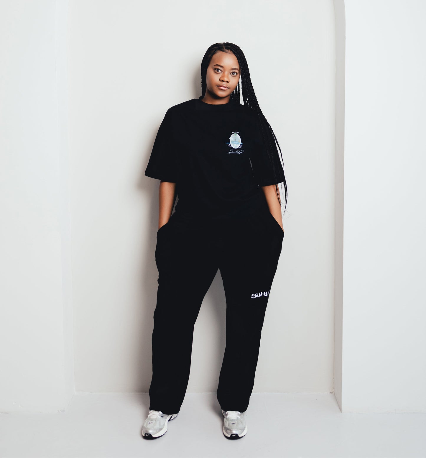 Model wearing a black SUHU  outfit with a logo, standing against a white wall.