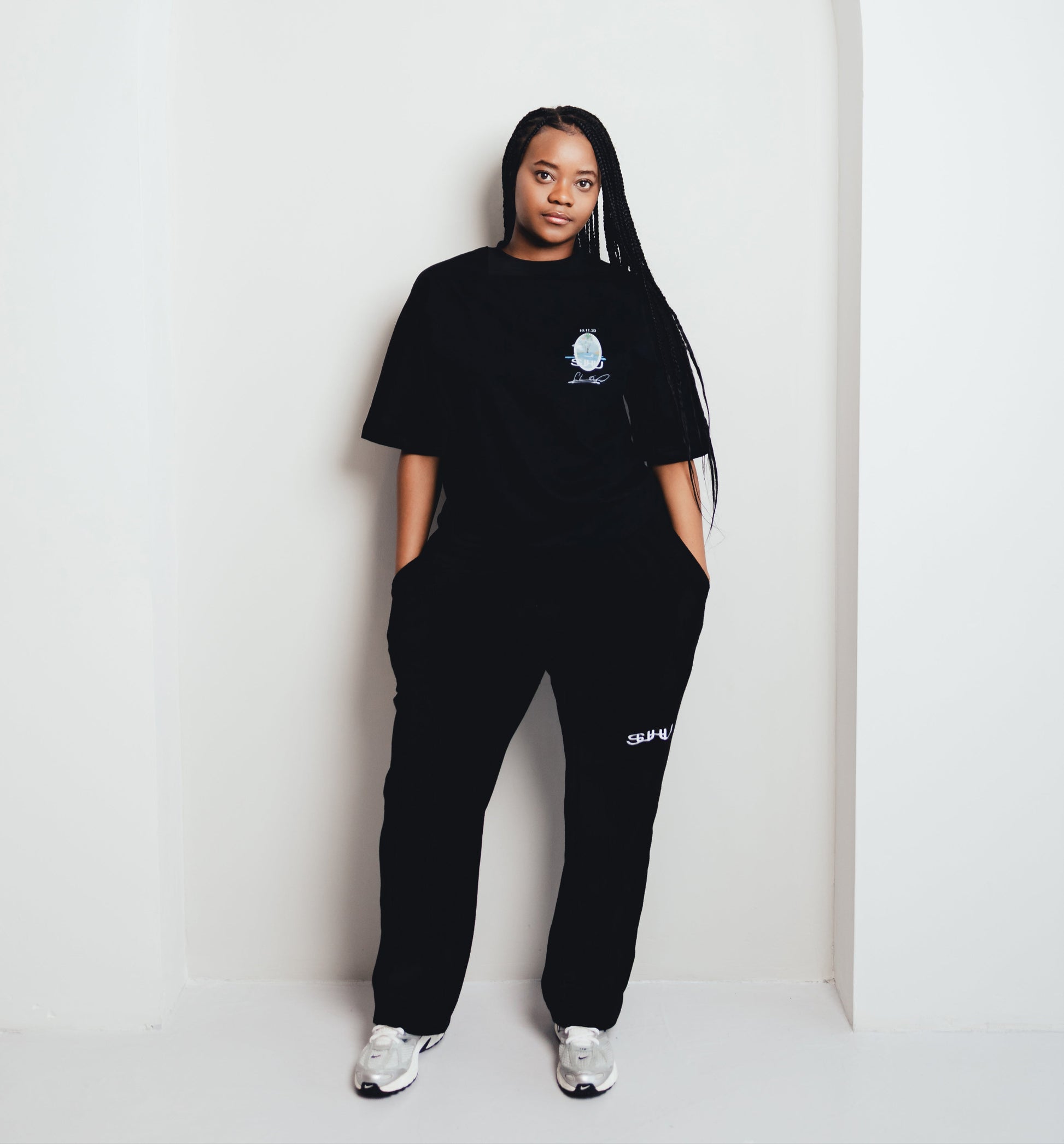 Model wearing a black SUHU  outfit with a logo, standing against a white wall.