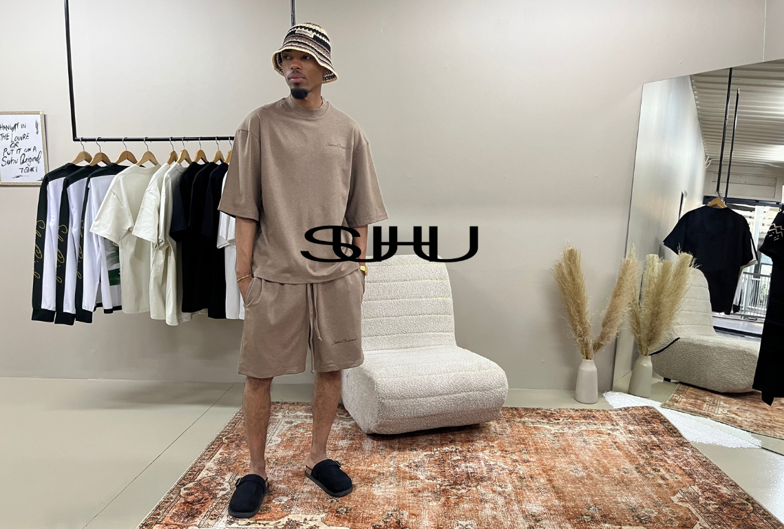 A SUHU model in a beige short-sleeve t-shirt and matching shorts set in a modern, stylishly decorated room with a mirror, clothing rack, and textured rug. The brand name 'SHU' is overlaid in the center.